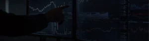 Person pointing at trading charts on multiple dark screens