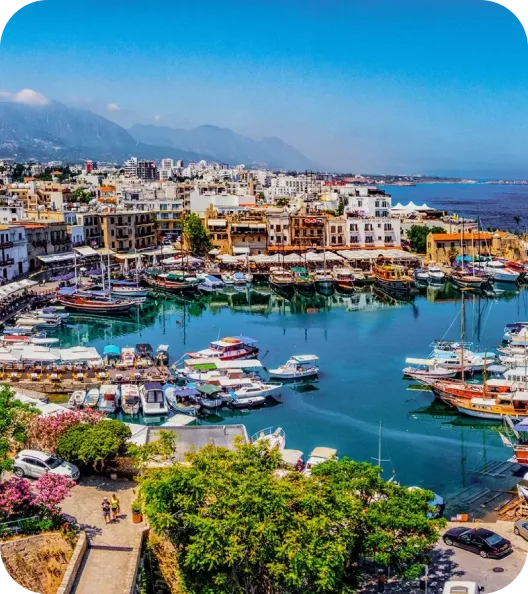 A scenic harbor with small boats and yachts, surrounded by old-style buildings and distant mountains under a clear blue sky.