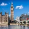 uk A tall clock tower beside a historic building and a bridge over a river on a sunny day with blue sky and clouds.